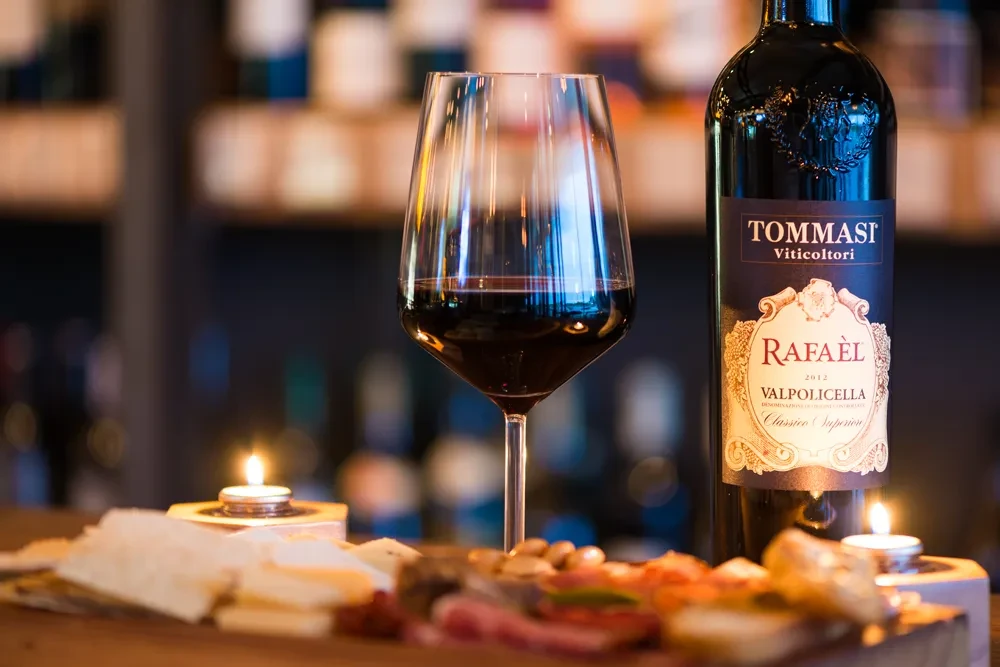 A glass of red wine, Tommasi bottle, charcuterie, and glowing candles.