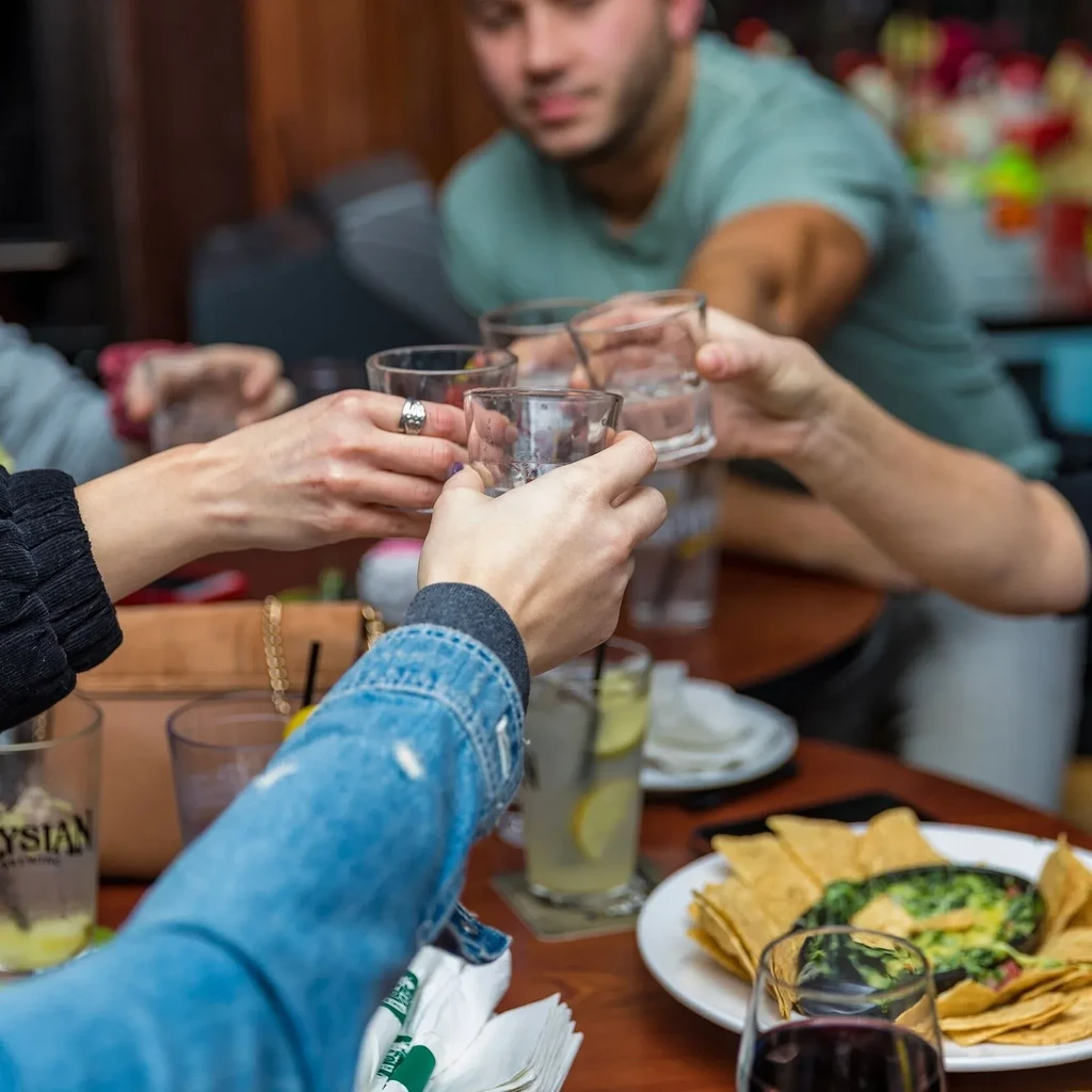 Multiple hands toast with clear glasses over chips and dip.