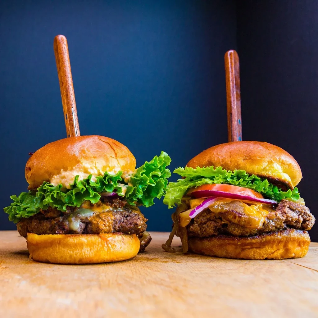 Two large beef burgers with cheese and fresh toppings on wood.