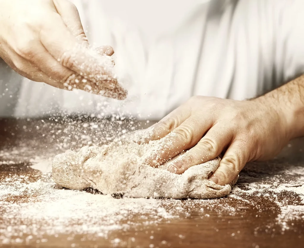 Close-up: Flour-dusted hands knead dough on wood, flour sprinkling.
