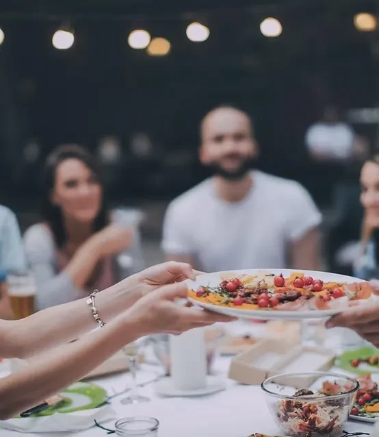 Hands pass a garnished dish at a blurred social dinner.