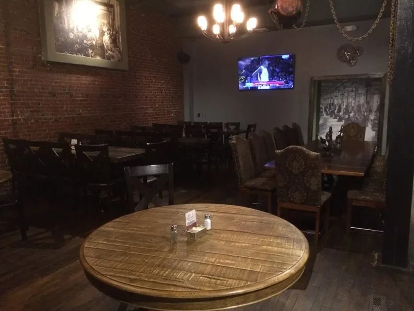 Dim, rustic restaurant interior with brick wall, TV, and wooden tables.