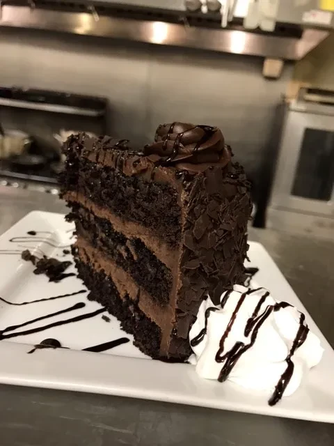 Multi-layered chocolate cake slice with whipped cream and sauce, professional kitchen.