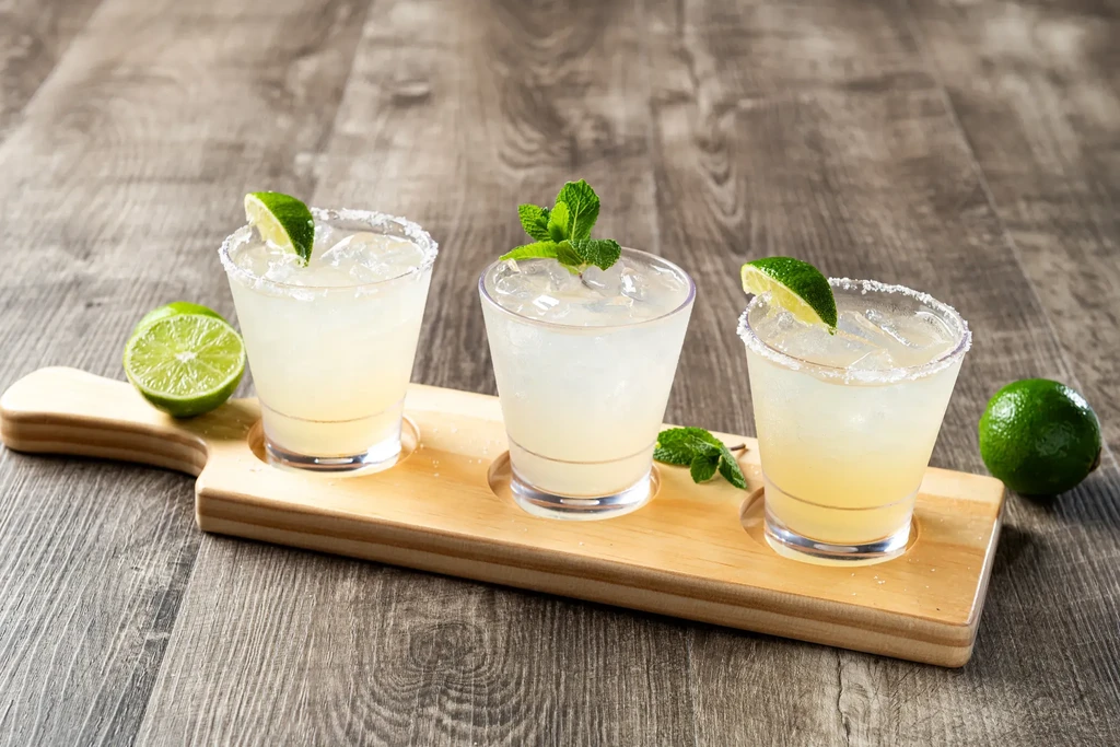 Three garnished cocktails with limes on a wooden flight board.