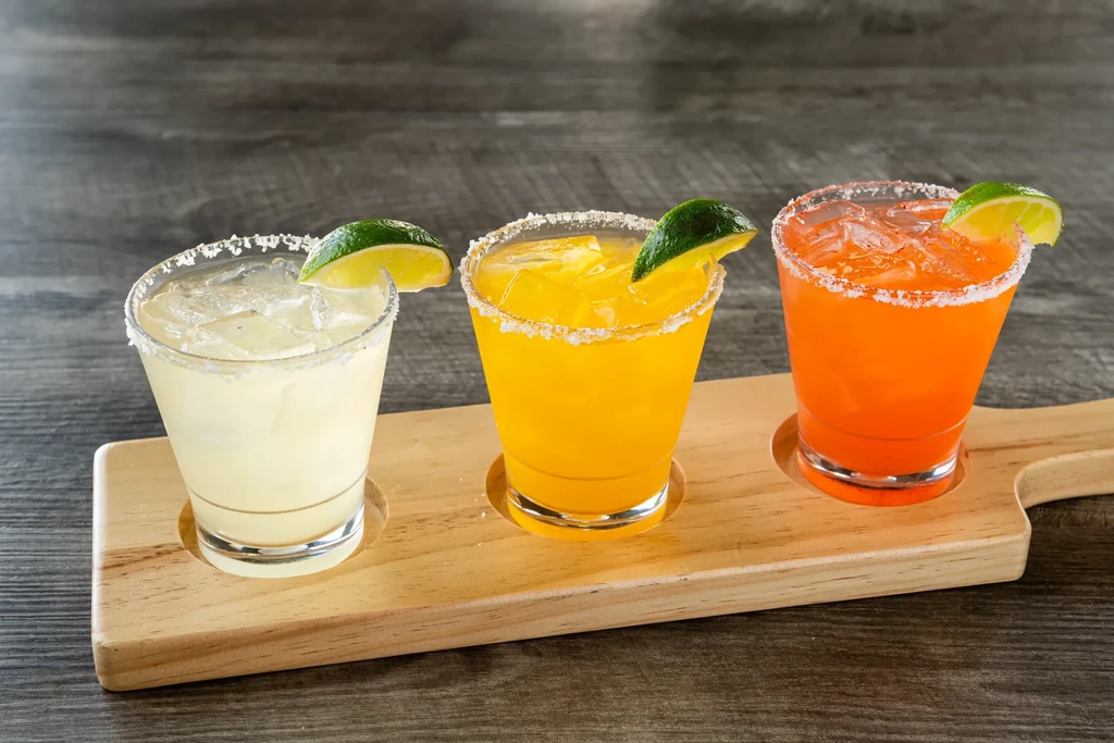 Three colorful salt-rimmed cocktails with lime on a wooden board.