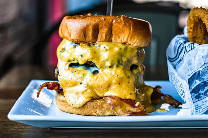 Multi-patty burger overflowing with cheese sauce, bacon, and onion rings.
