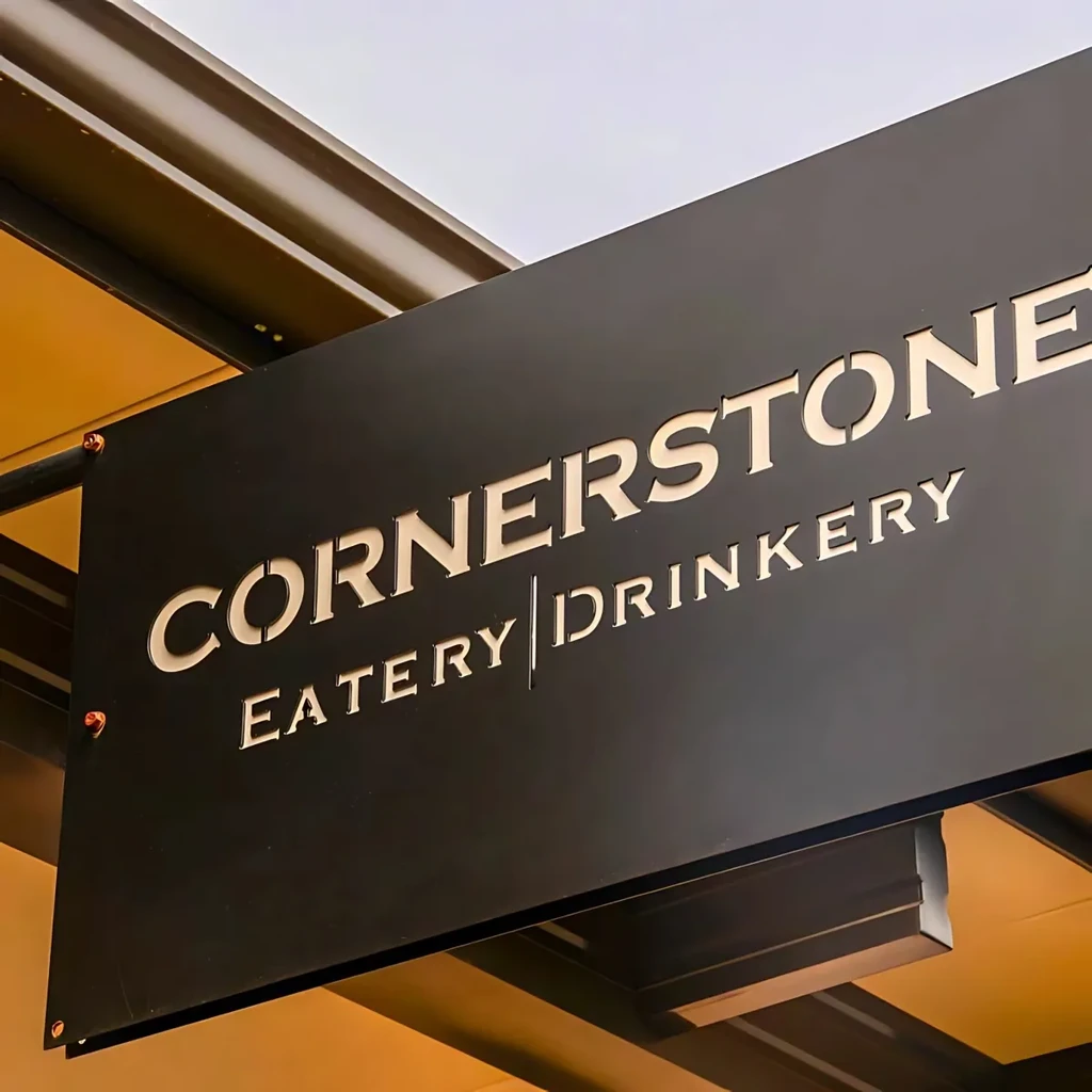 Black "CORNERSTONE EATERY | DRINKERY" sign, warmly lit, hanging on building.
