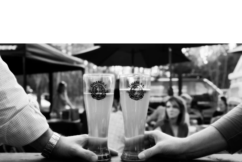Two hands hold Primal Brewery pilsners at an outdoor gathering.