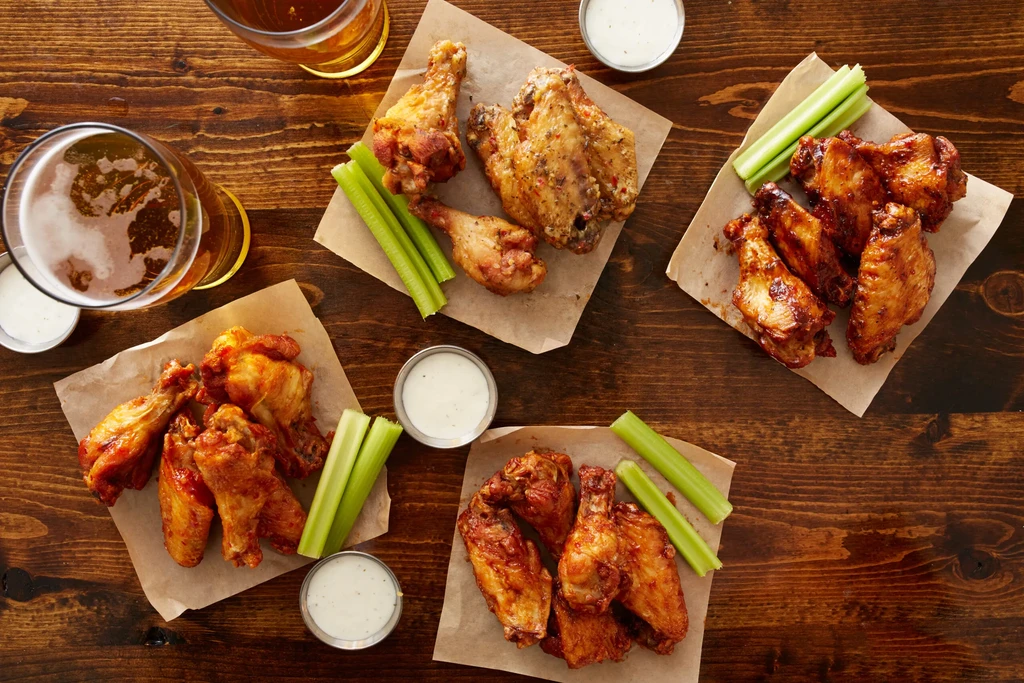 Four piles of chicken wings, celery, dips, and beer on a table.