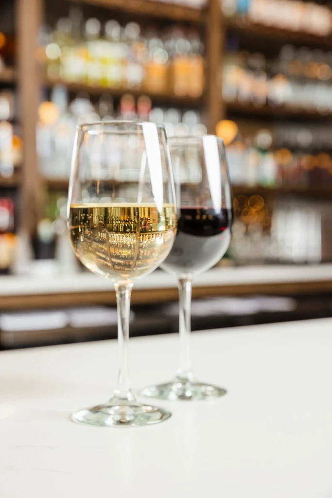Two wine glasses, white and red, on a bar with liquor bottles.