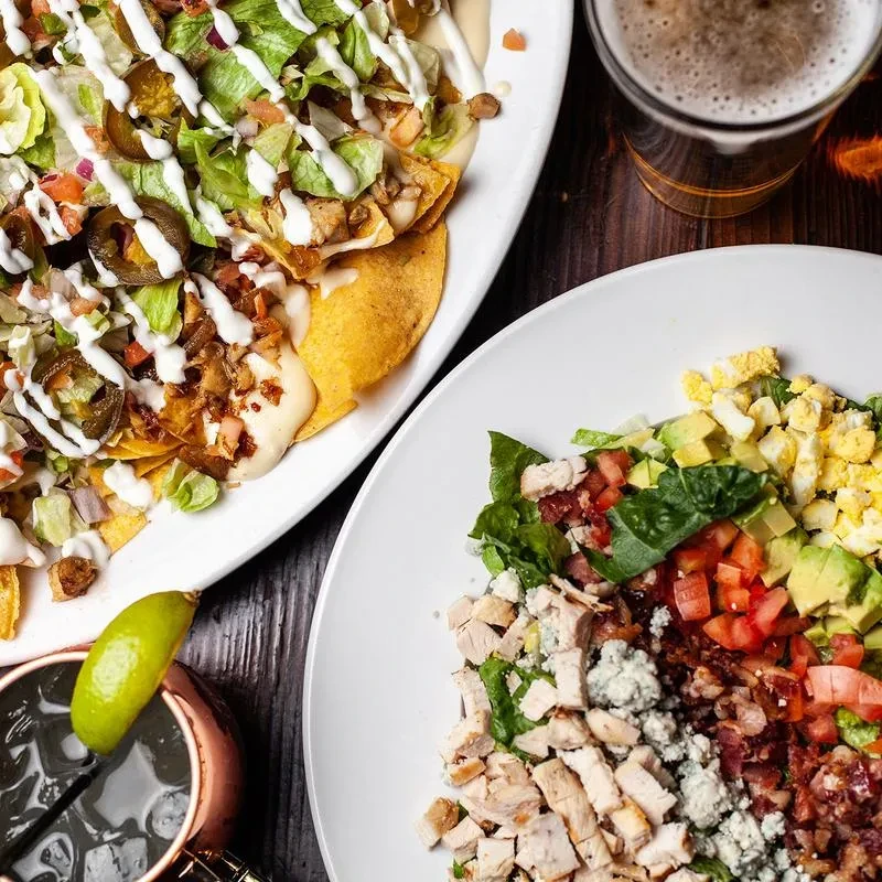 Overhead view: Nachos, Cobb salad, beer, Moscow Mule on a dark table.
