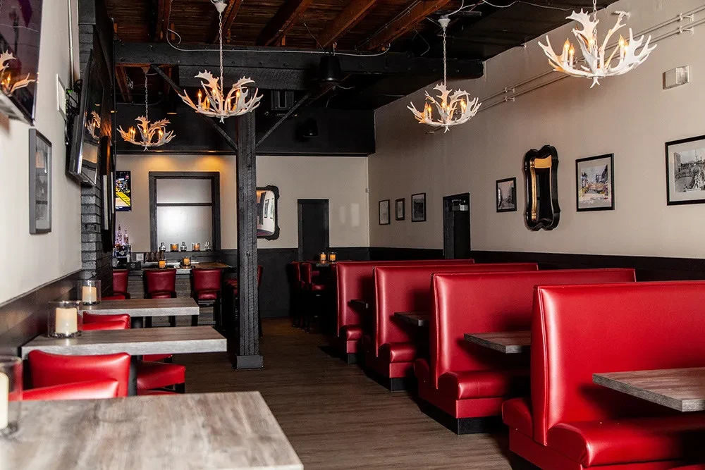 Modern rustic restaurant interior with red booths, antler chandeliers, and bar.