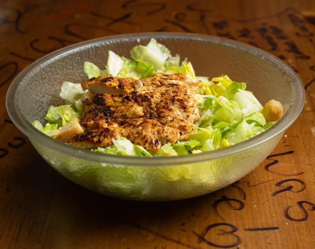 Grilled chicken Caesar salad in clear bowl on carved wooden surface.