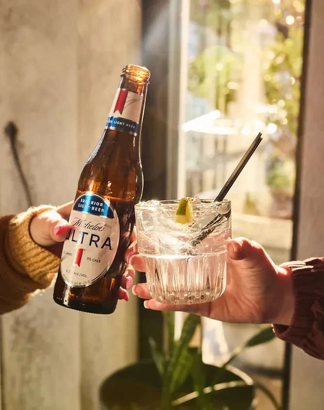 Two hands holding a Michelob Ultra beer and a clear drink with lime.