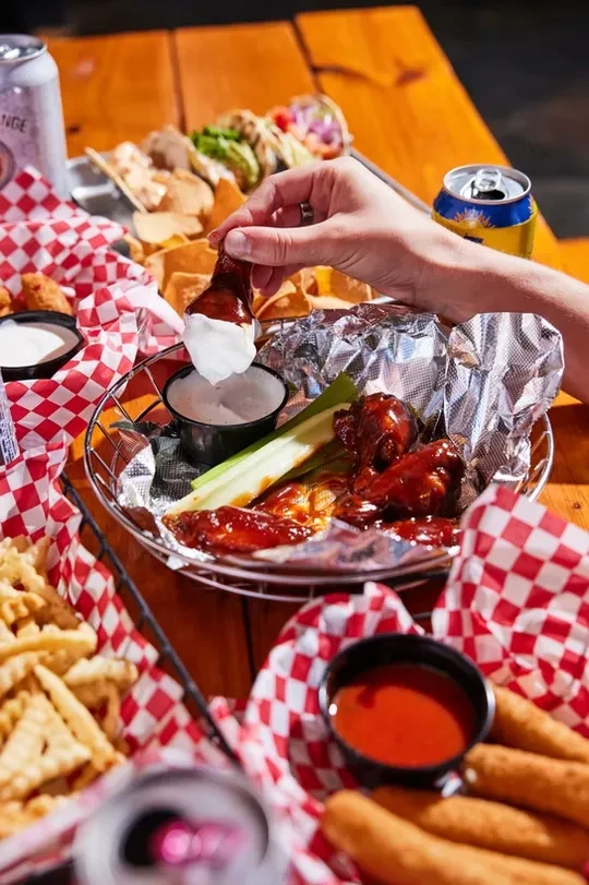 Hand dipping a chicken wing, with fries, mozzarella sticks, tacos.