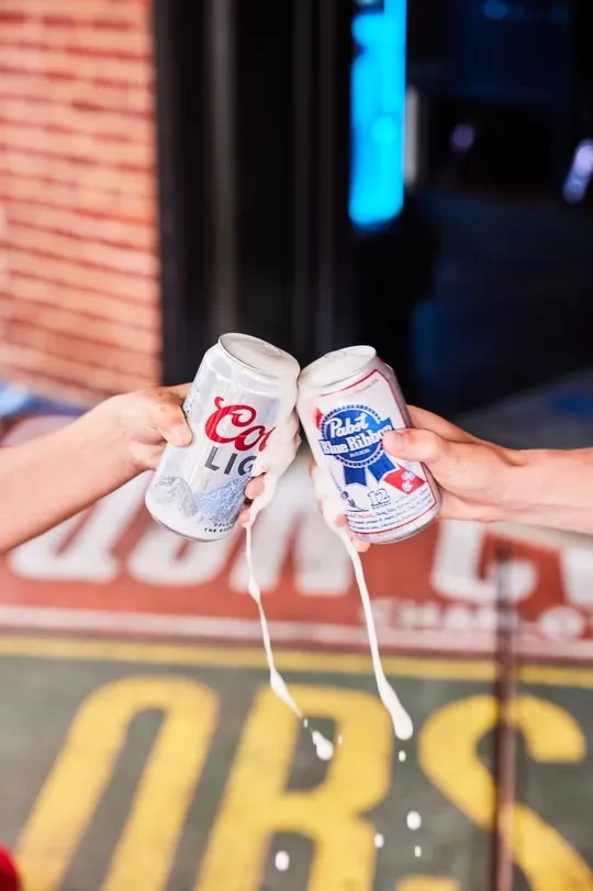 Hands spill Coors Light and Pabst Blue Ribbon cans.