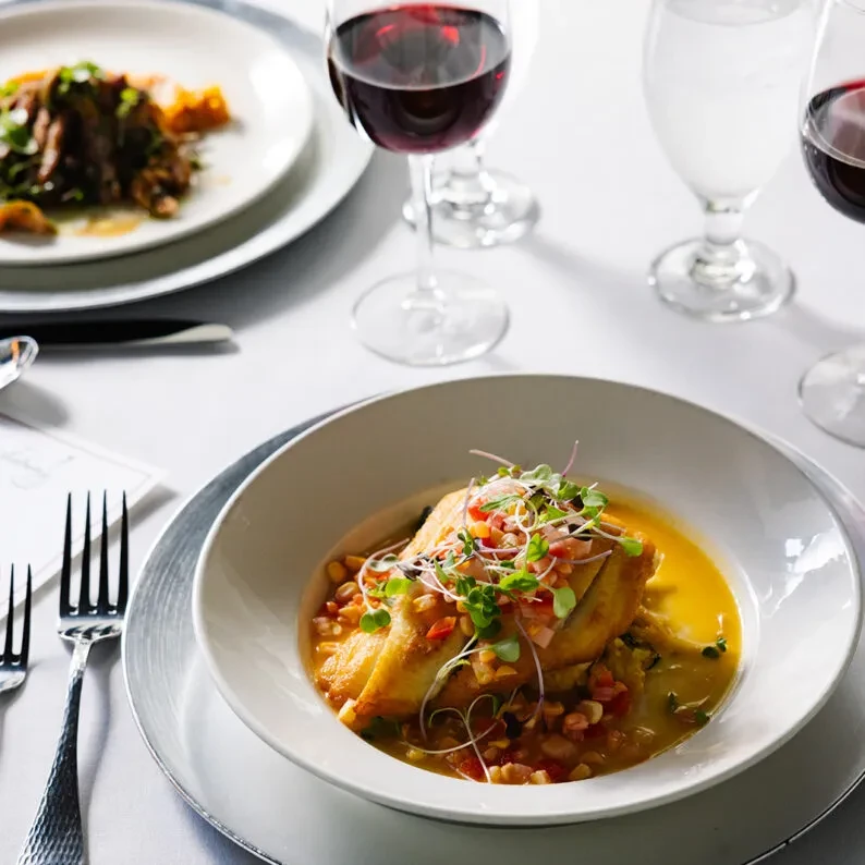 Close-up: Plated fish dish with sauce, wine glasses on elegant table.