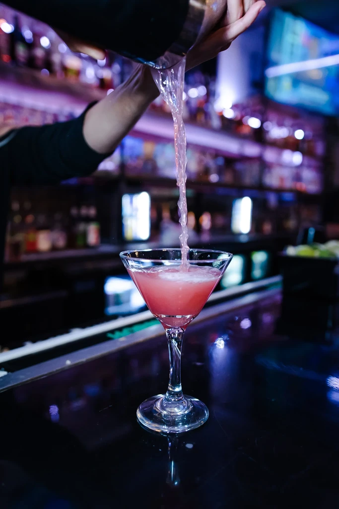 Bartender pours a pink cocktail from a shaker into a martini glass.