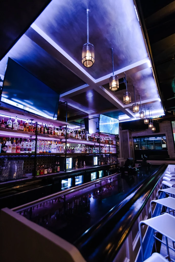 Well-stocked modern bar with purple-pink backlit liquor bottles and TVs.