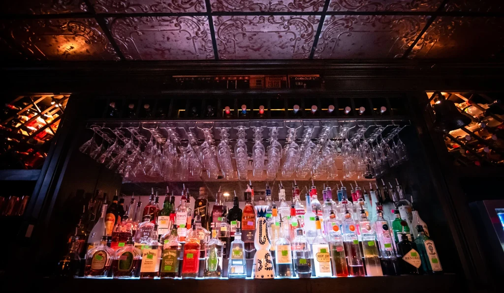 Dark bar with colorful liquor bottles, hanging glasses, and ornate ceiling.
