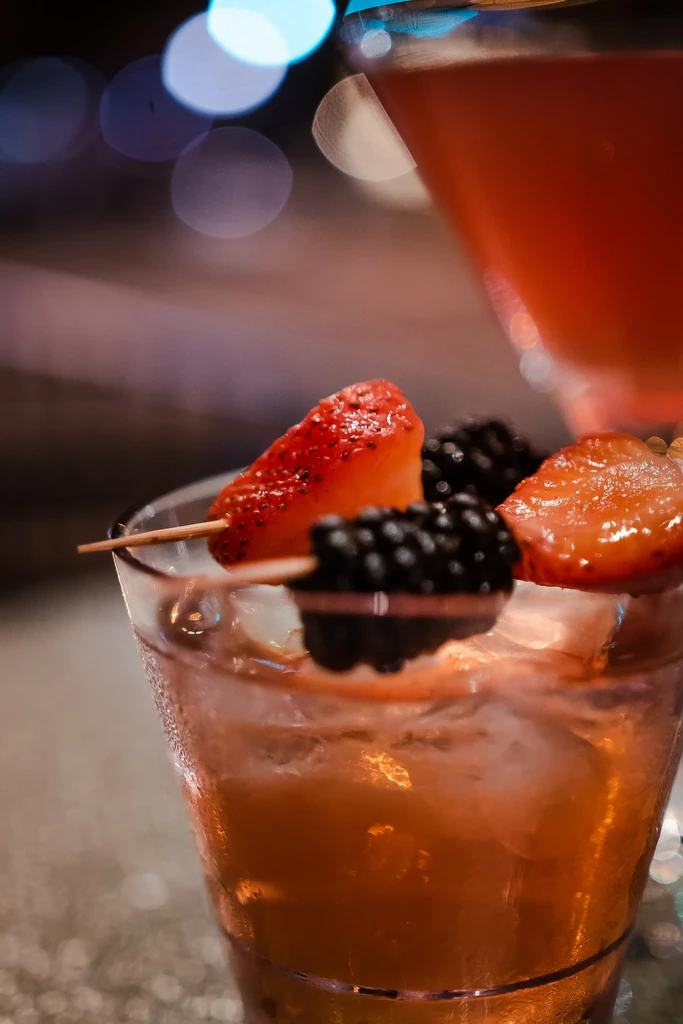 Amber cocktail with fruit skewer garnish, next to a blurred drink.