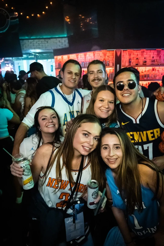 Eight young adults smile, holding drinks in a dim bar.