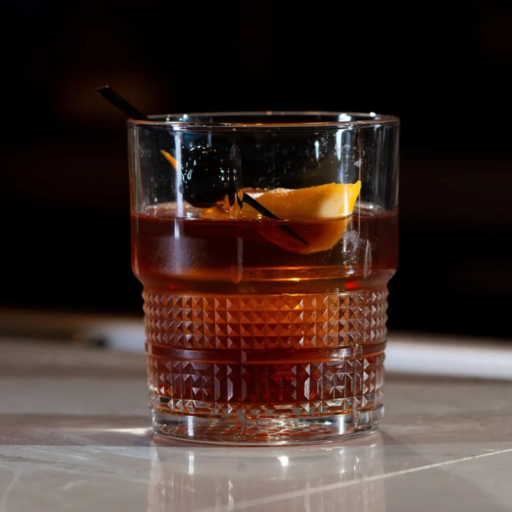 Amber Old Fashioned cocktail in a textured glass with cherry and orange.