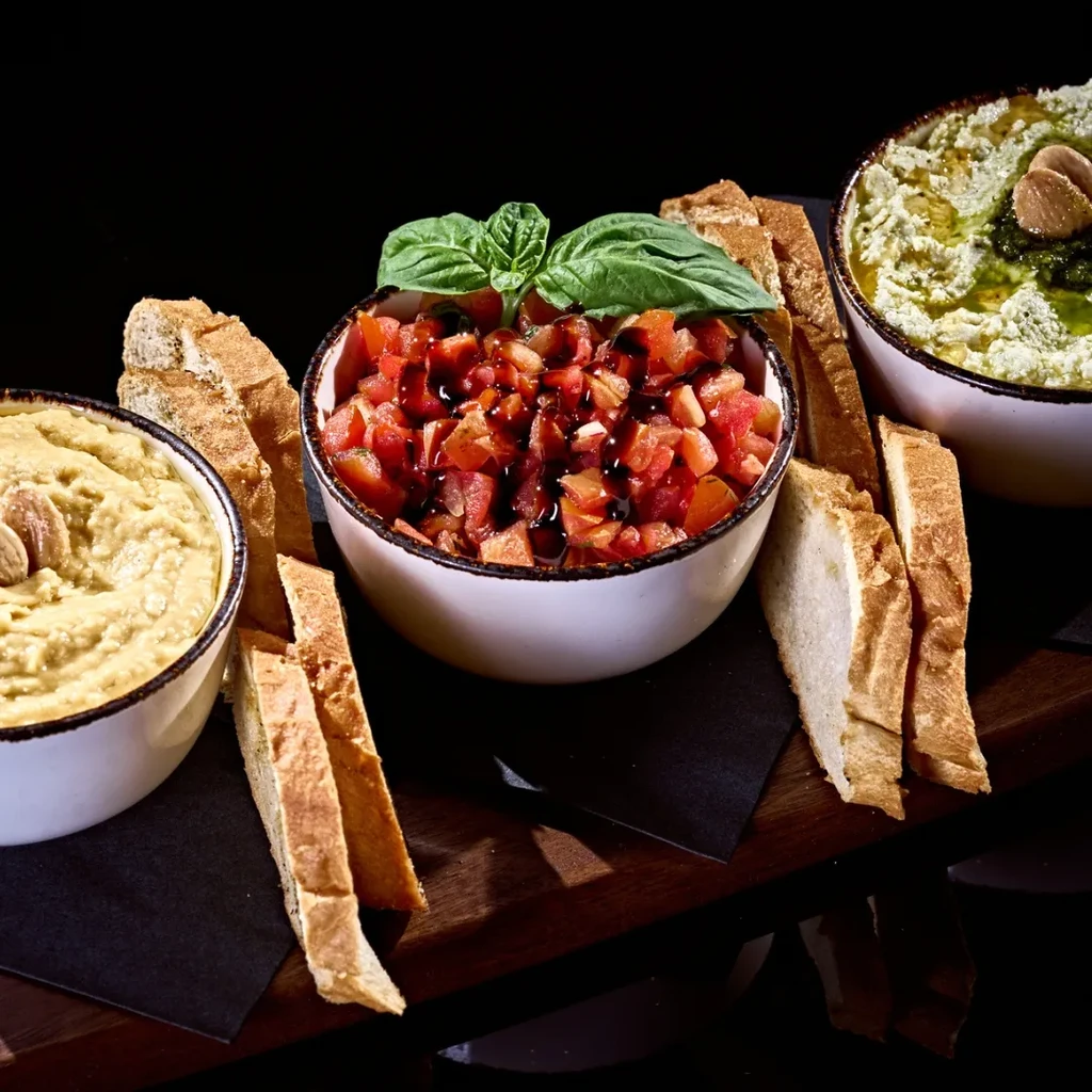 Appetizer board with bruschetta, hummus, green dip, and bread.
