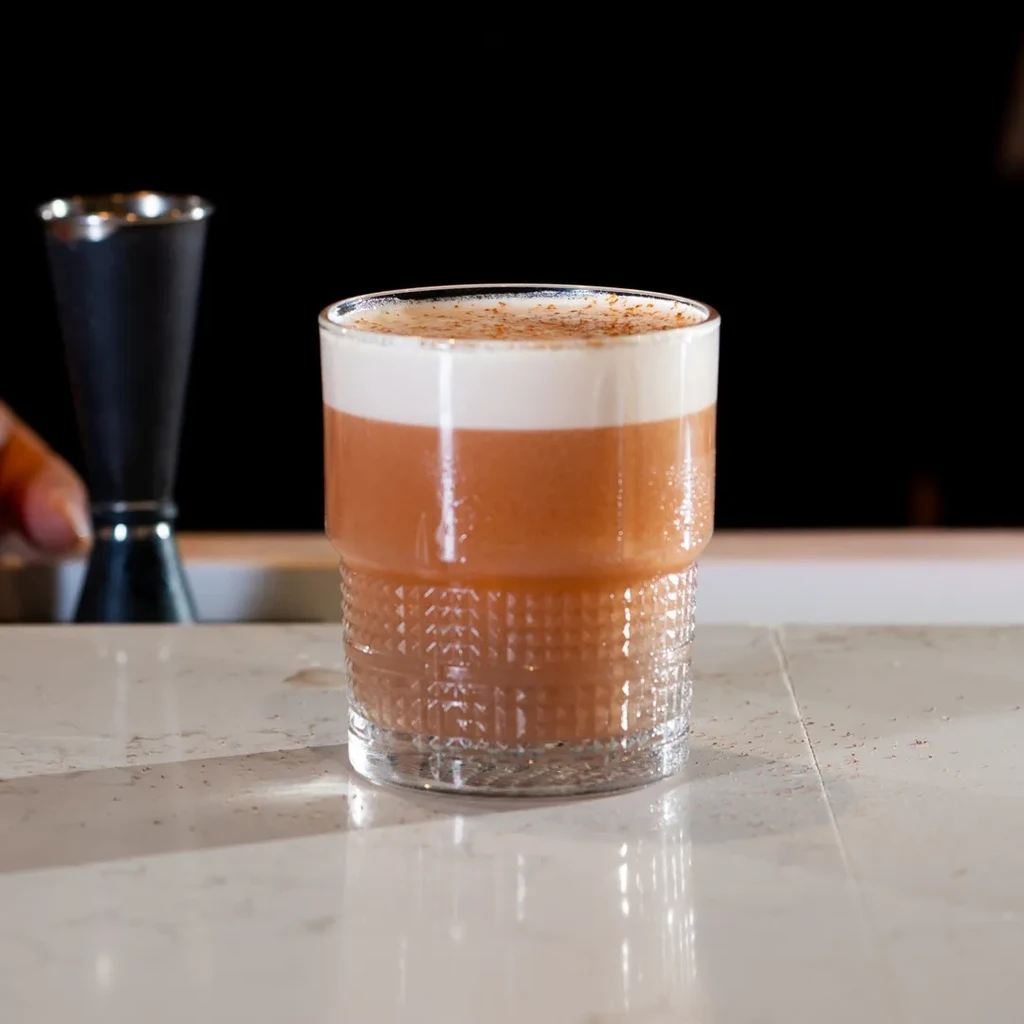 Caramel-colored drink with white foam and spice in textured glass.