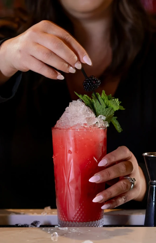 Hands garnish a red cocktail with crushed ice, mint, and a blackberry.