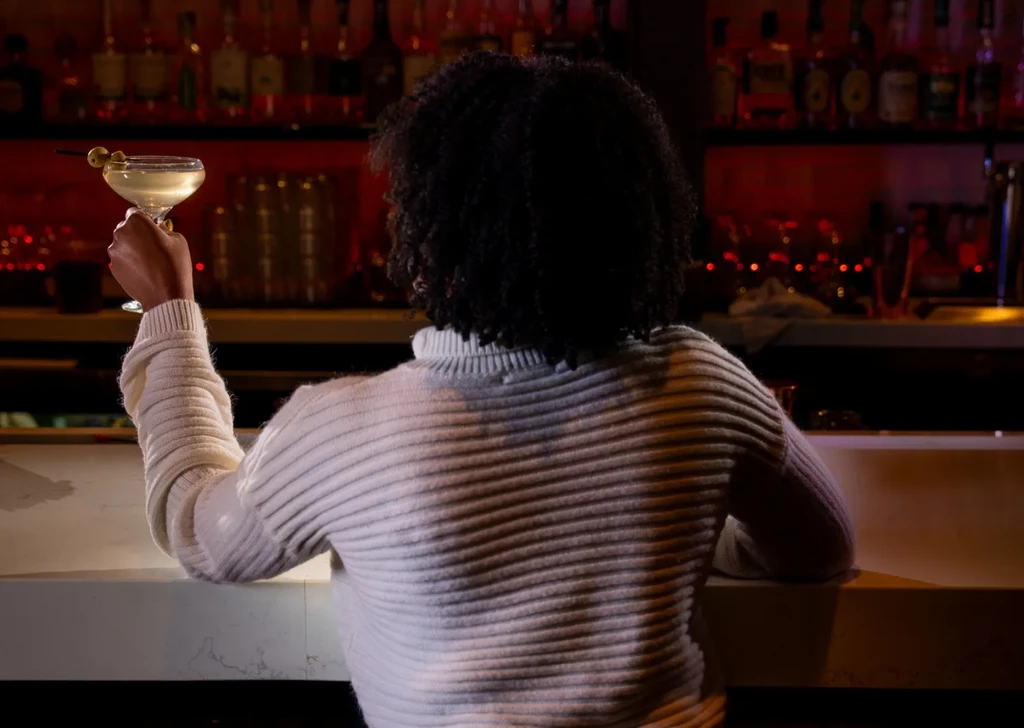 Woman from behind, holding an olive cocktail at a dimly lit bar.