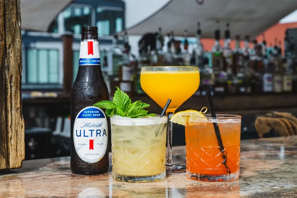 Michelob Ultra bottle and three colorful garnished cocktails on bar.