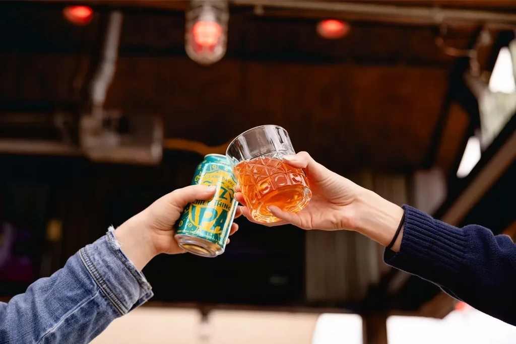 Two hands toast a beer can and glass of amber liquid.