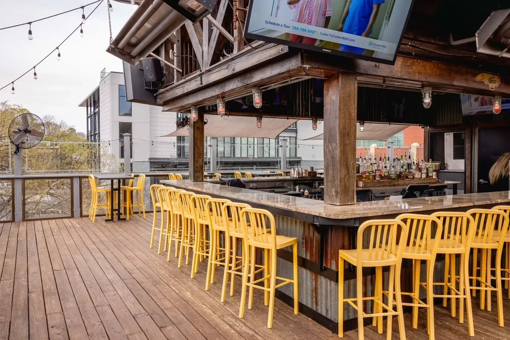 Outdoor rooftop bar with yellow stools, wooden deck, and city view.