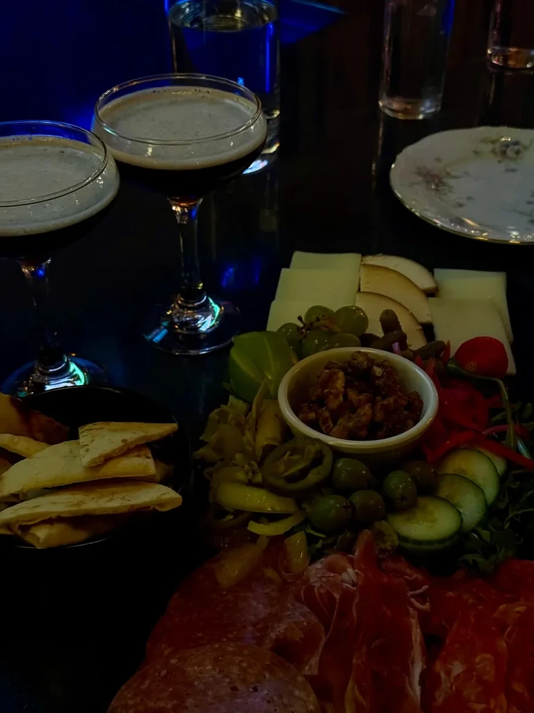 Charcuterie board, flatbread, and two dark cocktails in dim blue light.