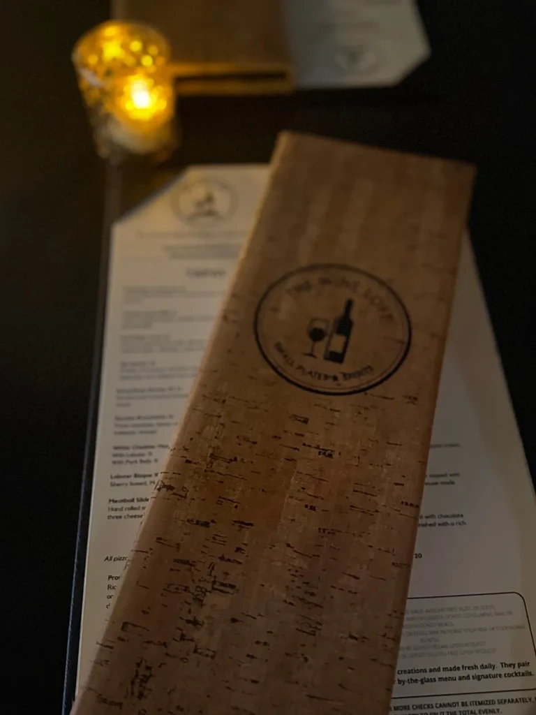 Rustic cork menu with "THE WINE LOUNGE" logo near a flickering candle.
