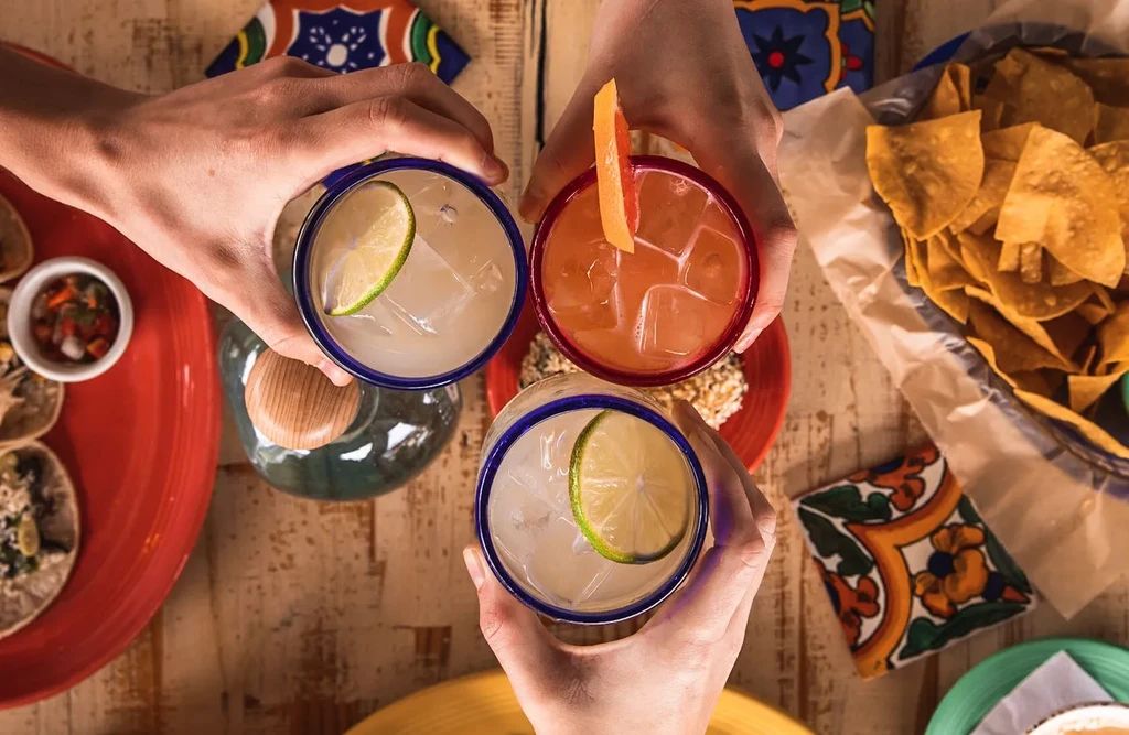 Three hands holding drinks, tacos, and chips on a table.