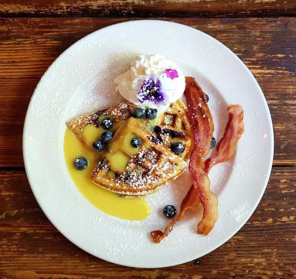 Overhead waffles, blueberries, bacon, whipped cream, yellow sauce on wood.