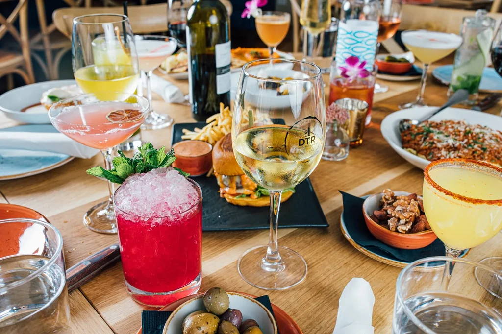 Dining table with colorful cocktails, wine, burgers, pasta, and snacks.