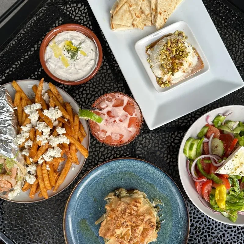 Overhead shot of Greek-inspired dishes and a cocktail on an outdoor table.