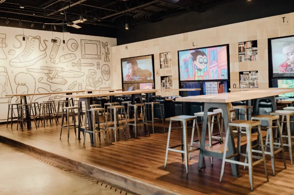 Industrial-chic restaurant with high-top seating, mural, and Scrabble-tile TV wall.