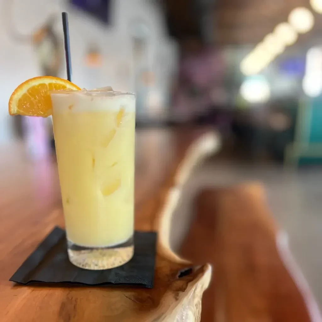 Yellow-orange drink with foam, ice, straw, and orange slice on wood.