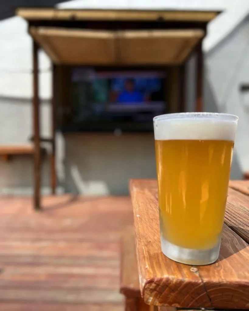 Glass of beer on a rustic table, blurred patio with TV.