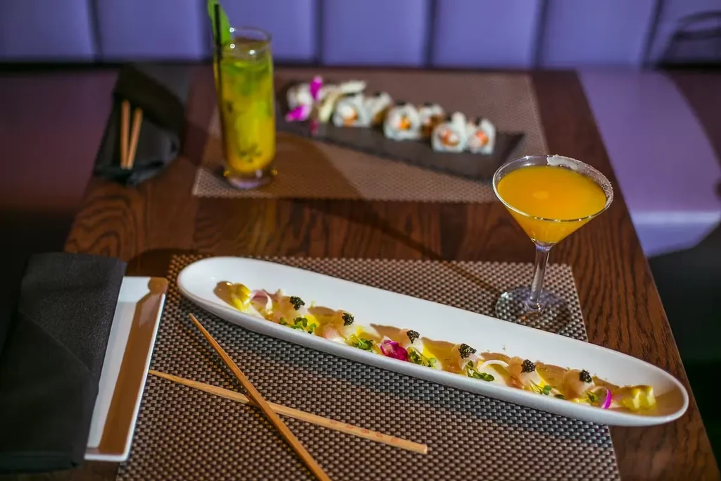 Upscale table with sashimi, cocktail, chopsticks, and sushi in blurred background.