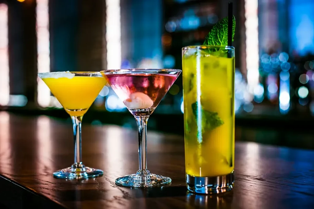 Three vibrant cocktails on a dark bar: yellow, pink lychee, green mint.