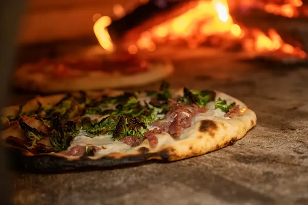 Pizza with meat and greens baking in wood-fired oven with flames.