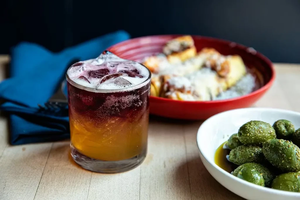 Close-up of a layered cocktail, green pickles, and food on wood.