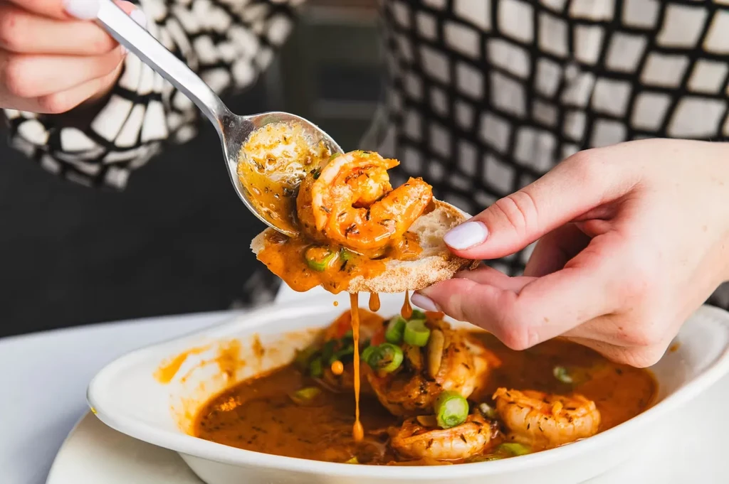 Hands dip toasted bread and spoon shrimp from a saucy bowl.
