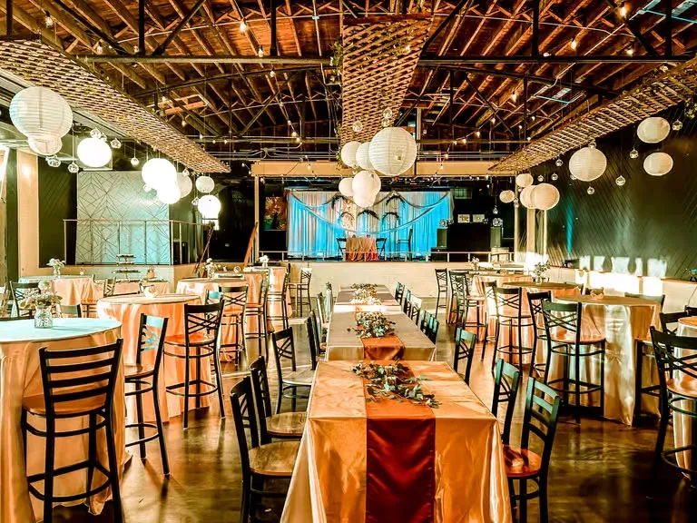 Warmly lit banquet hall with orange tables, flowers, and suspended lanterns.