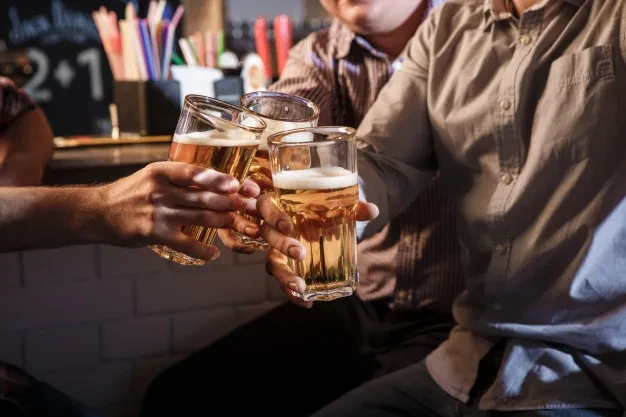 Three hands clink ribbed glasses of frothy amber beer at a bar.